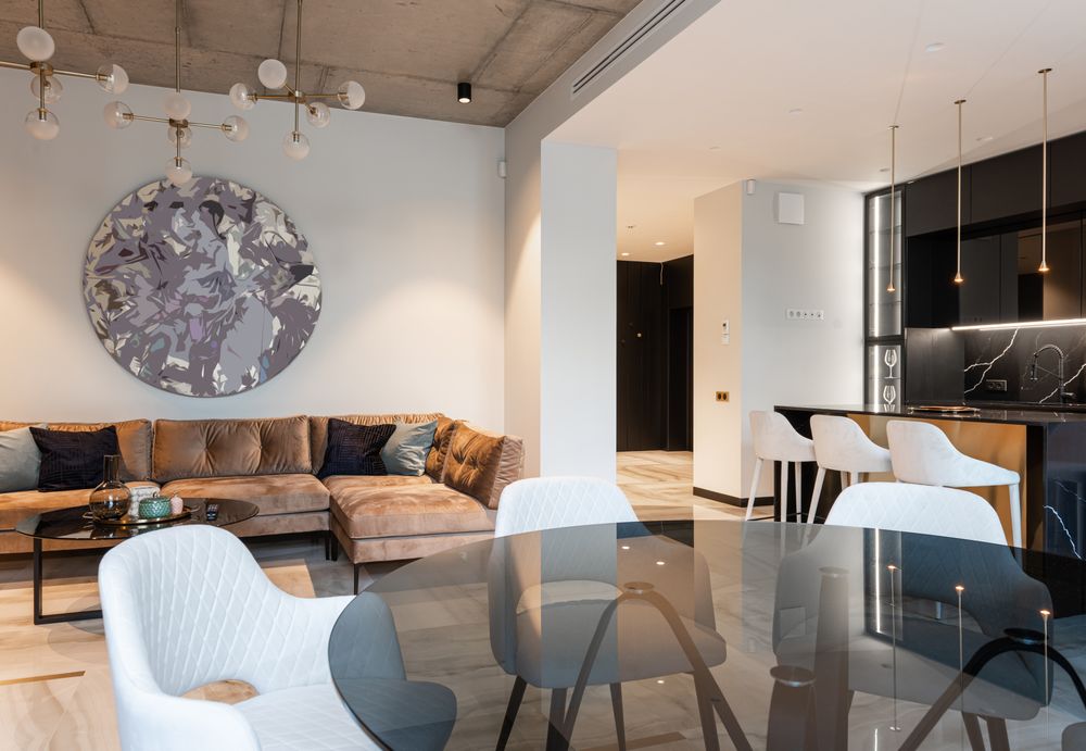 Stock photo: open-plan living room with a tan sectional sofa, large round wall art, a glass dining table with white chairs and a dark kitchen with bar stools.
