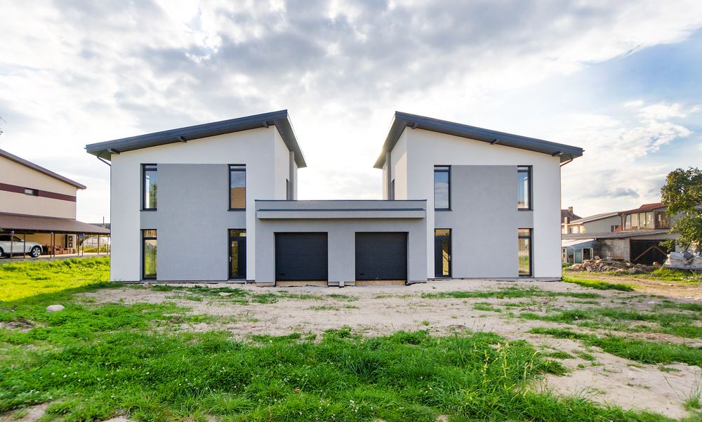 Stock photo: a pair of new two-storey white and grey houses with dark garage doors, on an unfinished grassy lot under a cloudy sky.